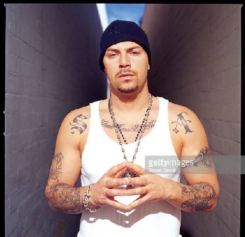 DJ Muggs standing with his white tank top and black cap showing his tattoos